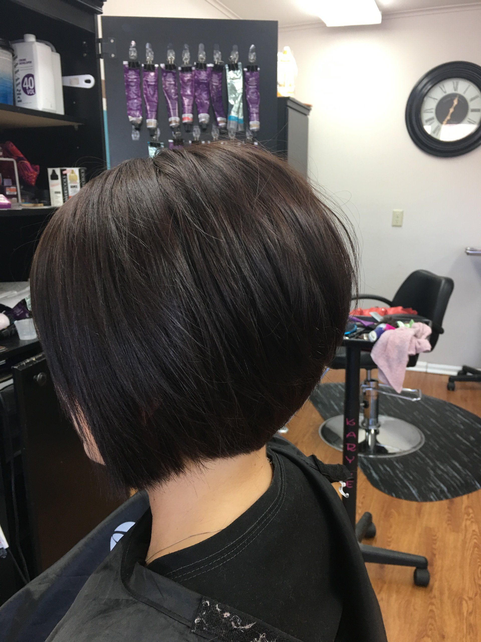 Short dark bob haircut in a salon chair, with purple product shelves and a wall clock in the background