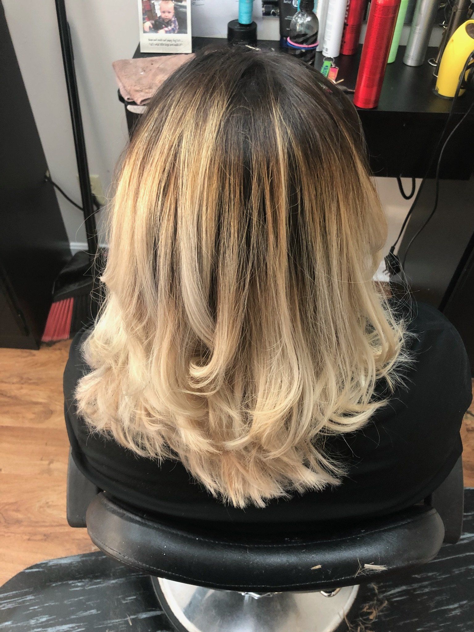 Back view of wavy blonde hair in a salon chair, with dark roots visible and shelves of products nearby