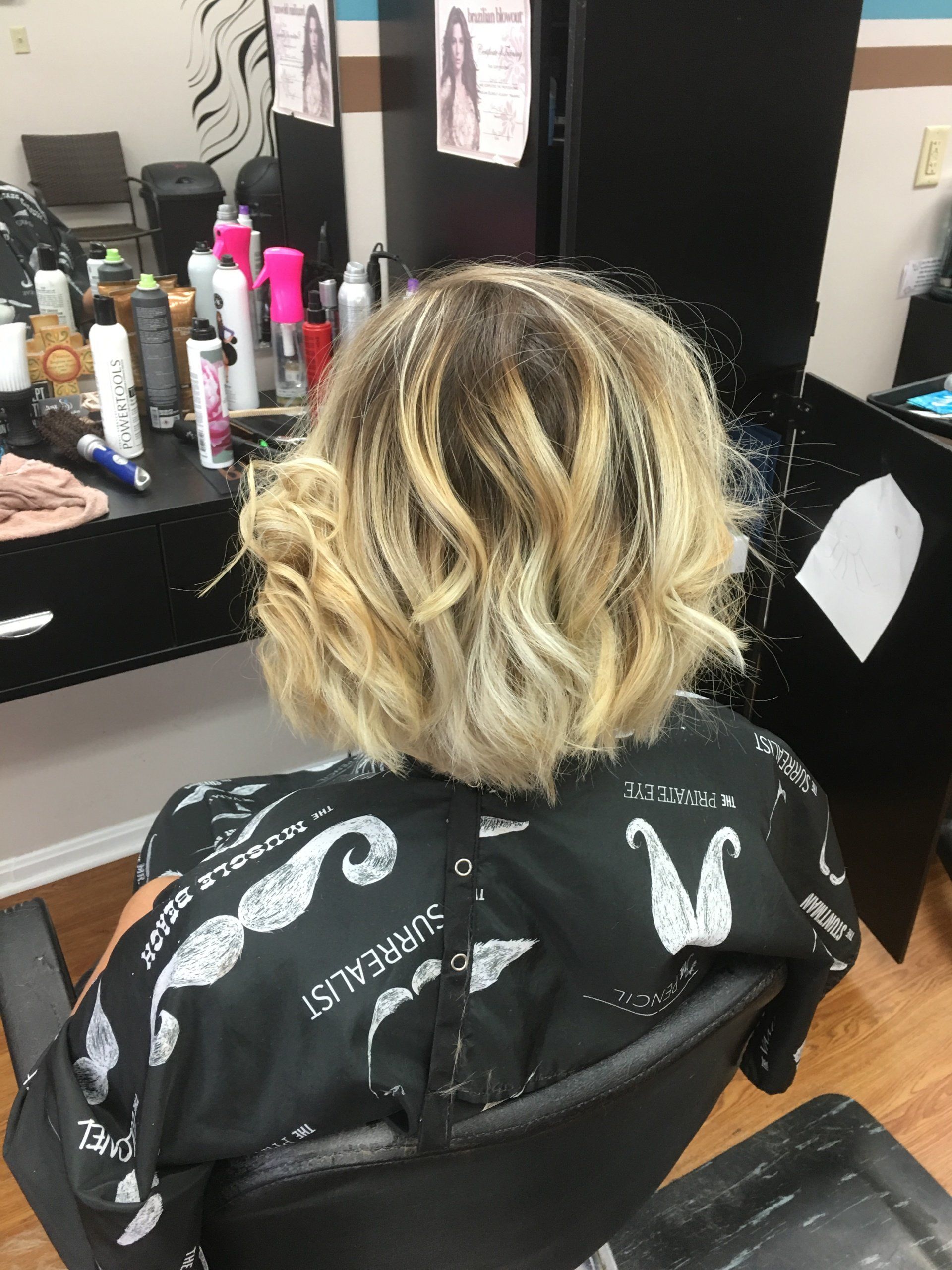 Person seated in a salon chair, seen from behind, with blonde curly hair and a black cape.
