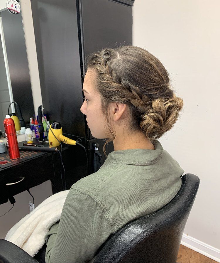 Person with braided bun sits in a salon chair, facing a mirror at a styling station with hair tools.