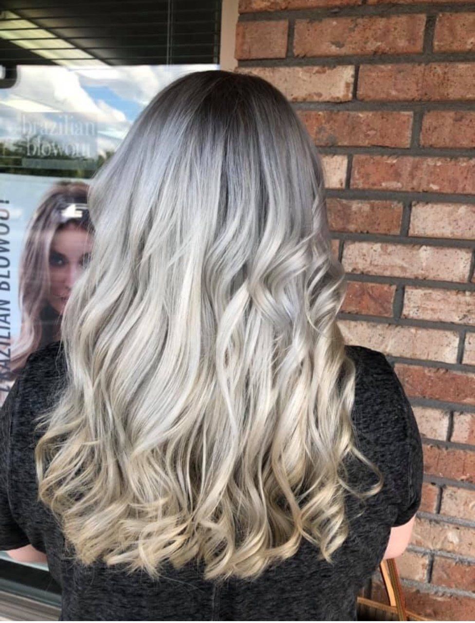 Long wavy silver-blonde hair with dark roots, seen from behind against a brick wall.