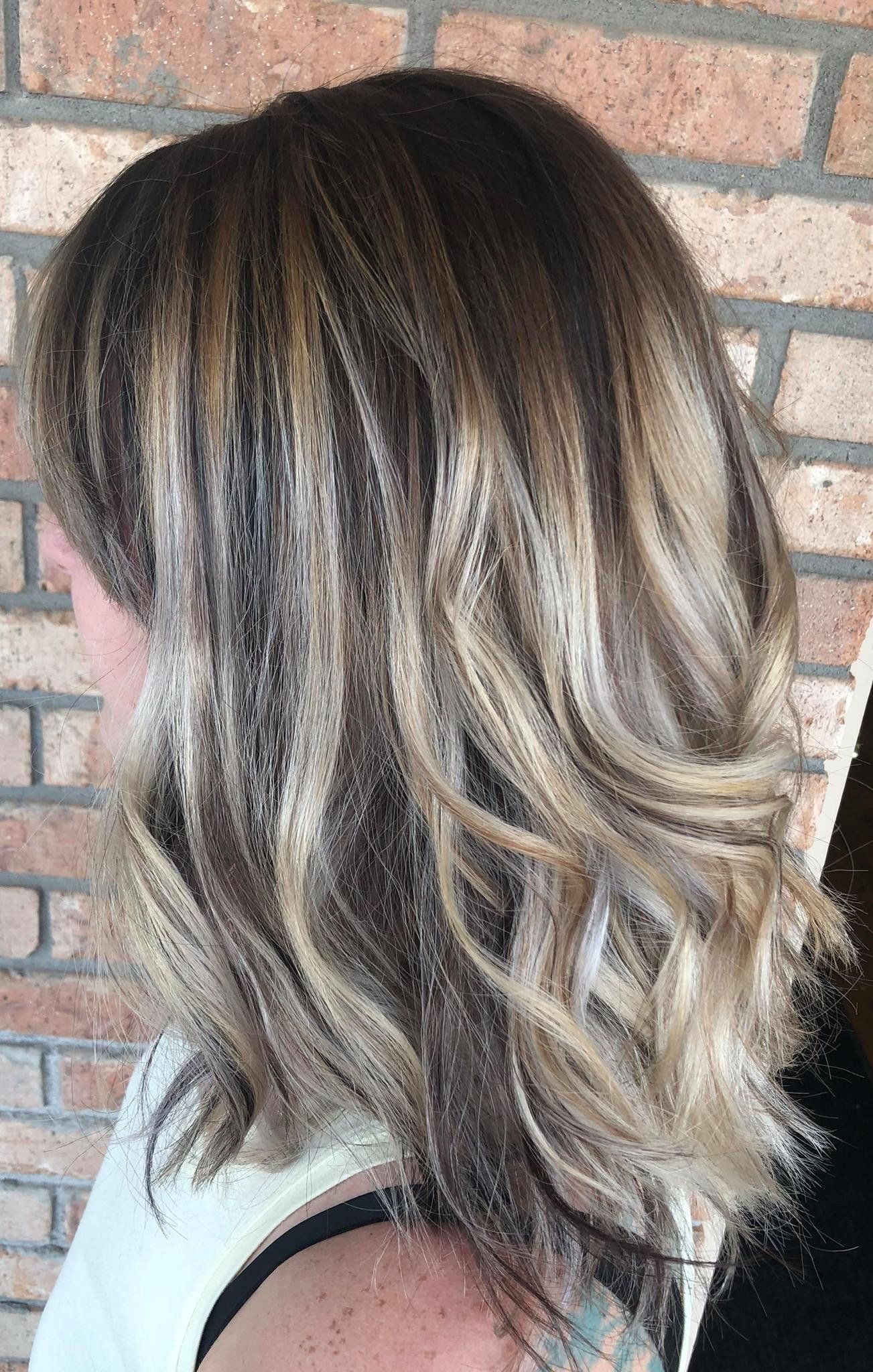 Side view of shoulder-length wavy hair with dark roots and blonde highlights against a brick wall