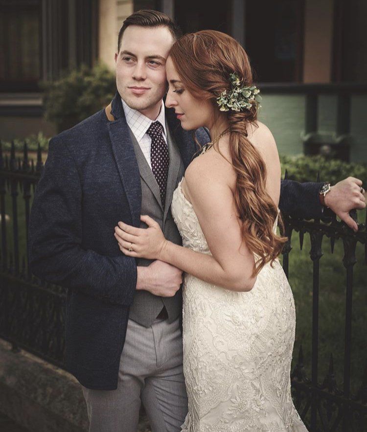 Couple embracing outdoors in formal attire, with the woman holding the man's arm and flowers in her hair