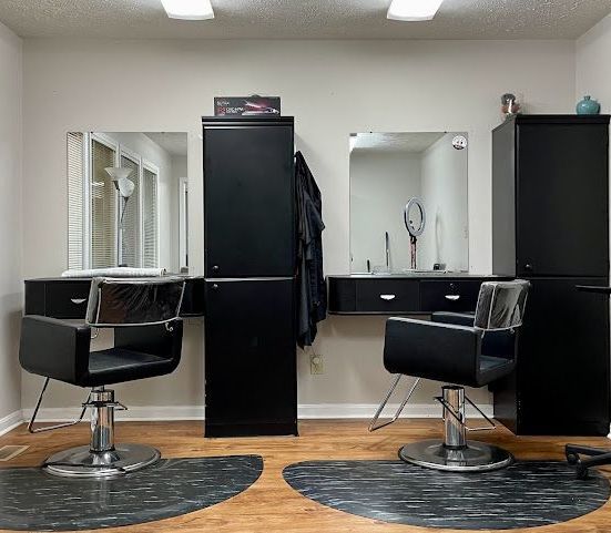 Hair salon interior with two black styling chairs, mirrors, and tall black cabinets on a wood floor.