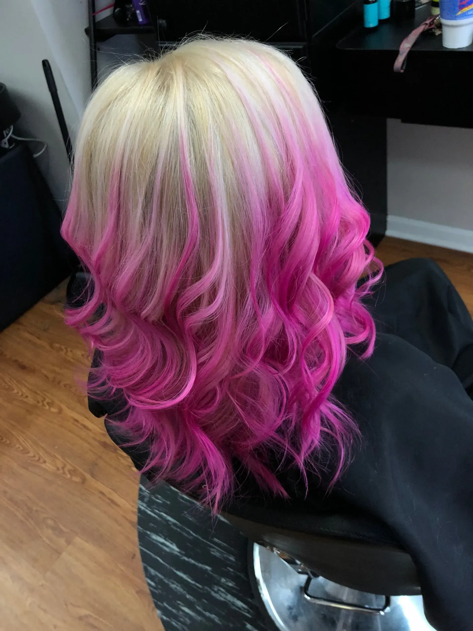 Blonde hair with bright pink curls, styled in soft waves over a black salon cape.