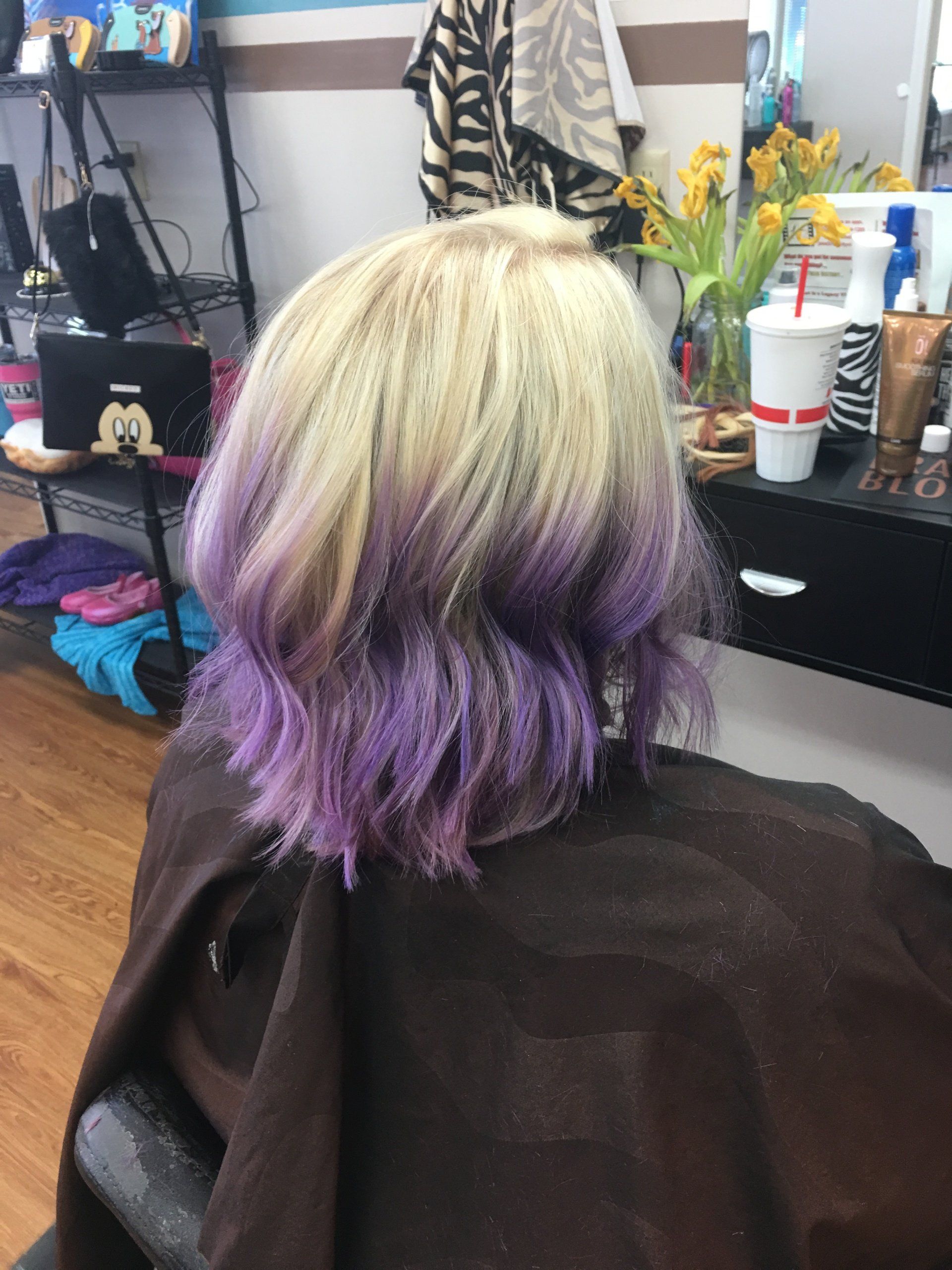 Blonde hair with purple ombré ends in a salon chair, with a mirror, flowers, and drinks in the background