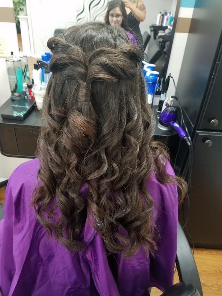 Back view of curled dark hair styled half-up with a braided twist, in a salon chair with purple cape