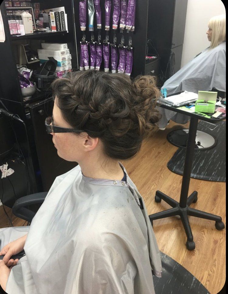 Person with braided bun sits in a salon chair, wearing a cape, as a stylist works nearby.