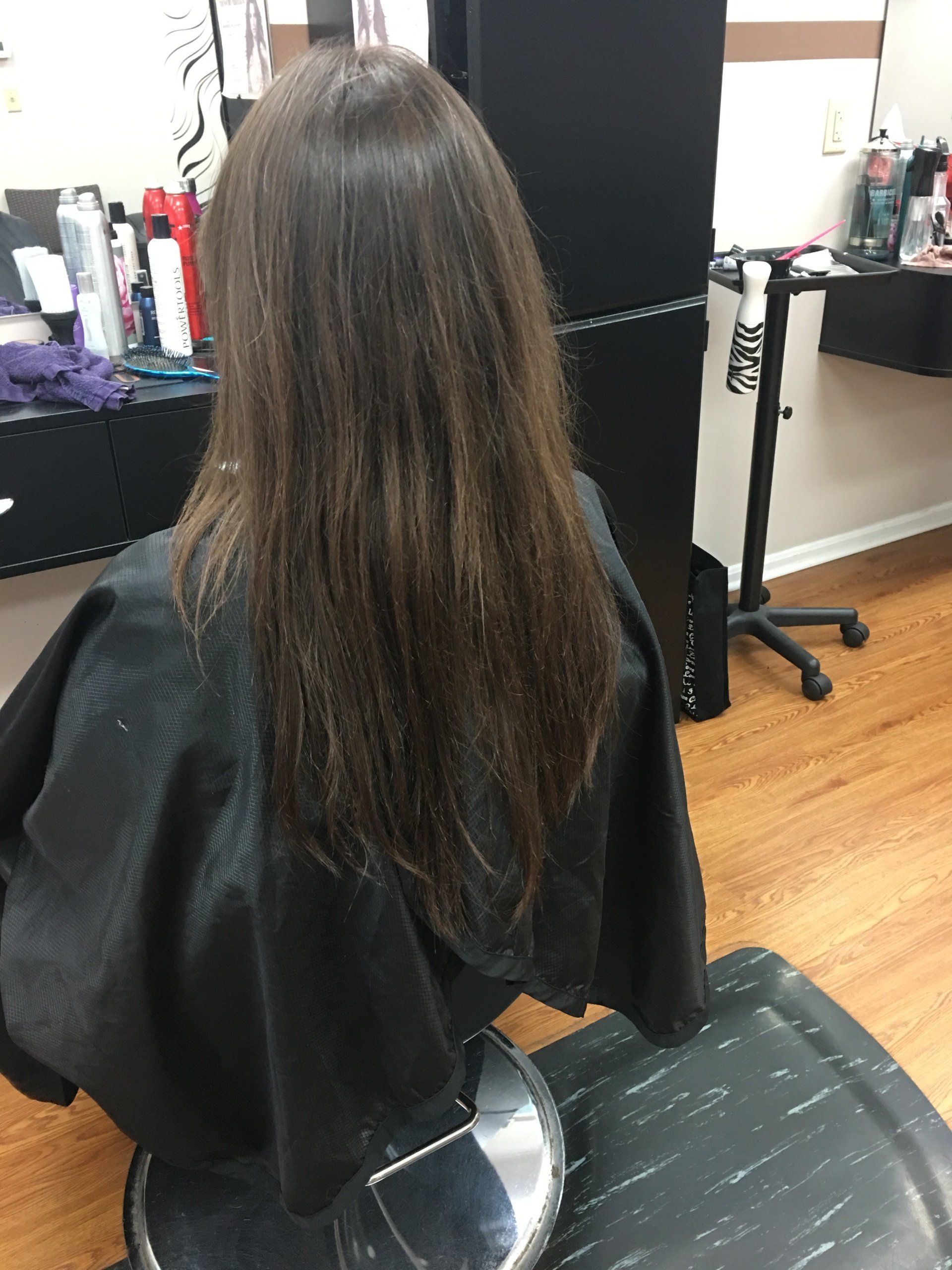 Person with long brown hair seated in a salon chair, viewed from behind, wearing a black cape.