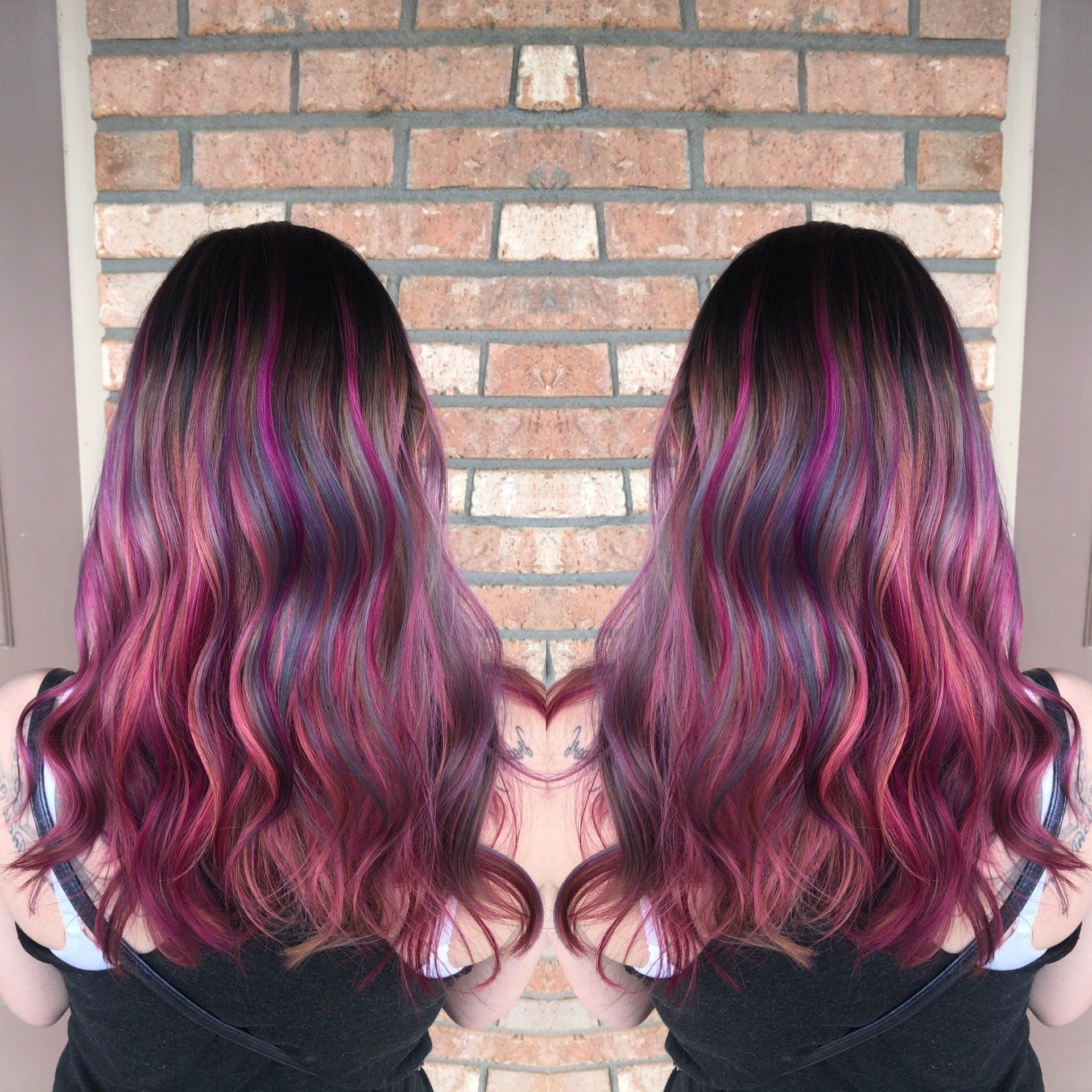 Back view of mirrored wavy hair dyed black with magenta highlights against a brick wall